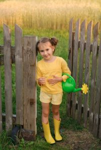 About Fence Sales of Sycamore. Young girl holding a green watering can and standing in a yard next to a fence.